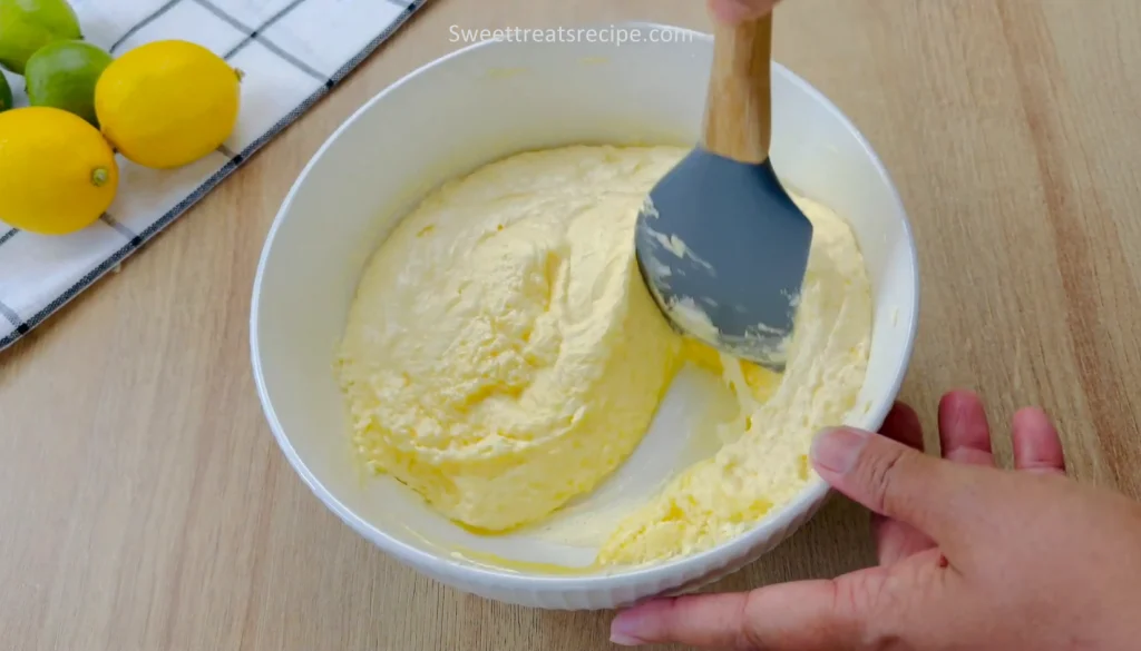 ixing creamy cake batter with a blue spatula in a white bowl, with lemons and limes in the background on a striped towel