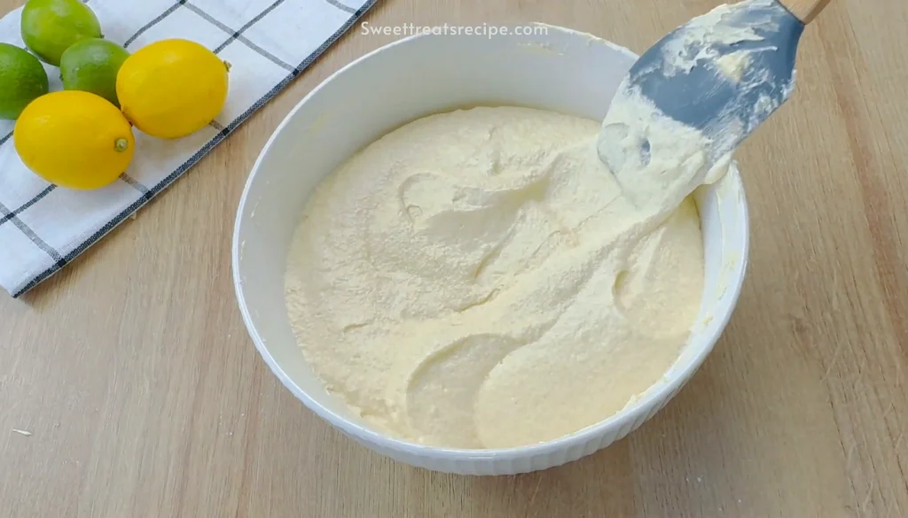 Mixing smooth cake batter in a white bowl with a blue spatula, while lemons and limes rest on a kitchen towel in the background.