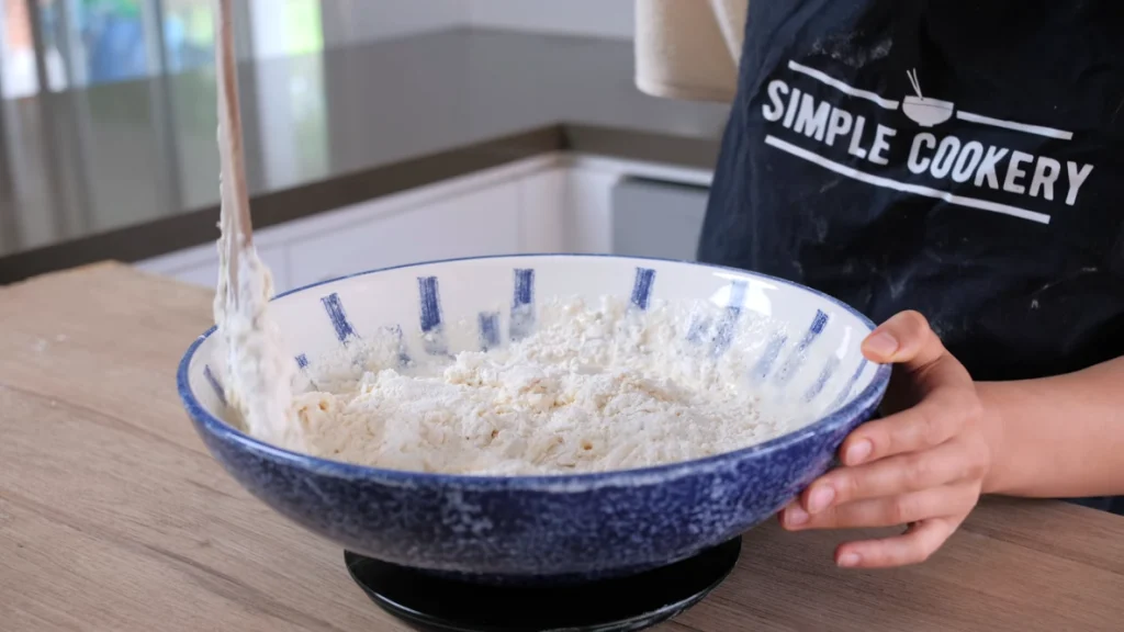 A bowl containing self-raising flour, thickened cream, and lemonade being mixed with a spoon until just combined. The dough is soft and sticky.