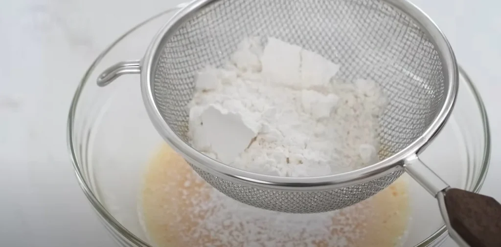 Cake flour being sifted through a fine mesh sieve into chiffon cake batter.