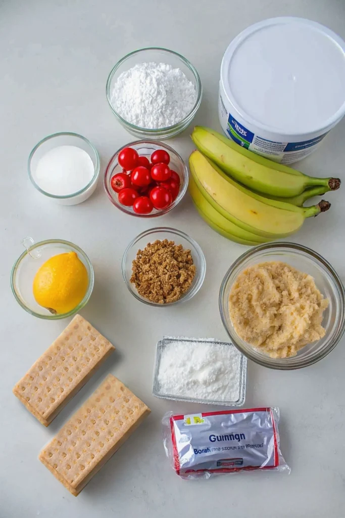 Overhead view of ingredients for No-Bake Banana Split Cake: bananas, maraschino cherries, graham crackers, powdered sugar, cream cheese, butter, and sugar arranged on a light surface.